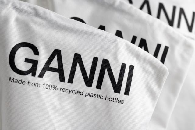 Ein Stapel von Textilien mit dem Markennamen 'GANNI' in großen schwarzen Buchstaben. Unter dem Namen steht der Slogan 'Hergestellt aus 100% recycelten Plastikflaschen' in kleinerer Schrift