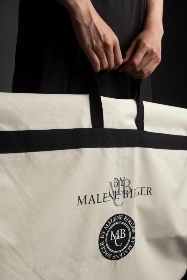 Person hält eine große Kleidersäcke, die hauptsächlich cremefarben mit schwarzen Akzenten am Rand und den Henkeln ist. Auf der Tasche ist ein Logo und der Name 'BY MALENE BIRGER